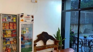 A room with a wooden bench, a small table, a refrigerator stocked with snacks, and a large window offering a view of the outdoors. - Sipalay Cozy Garden Stay (Sipalay)