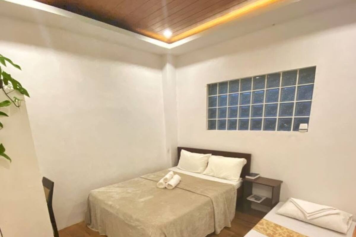 A small, well-lit room with a wooden ceiling, a bed with a beige bedspread, a bedside table with a lamp, and a window with blue-tinted glass.