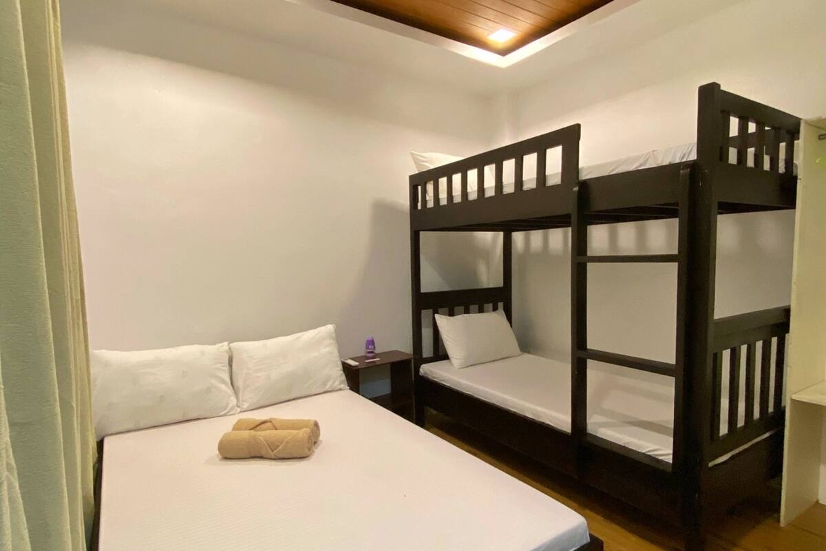A room with a bunk bed, a single bed, a small table, and a wooden ceiling.