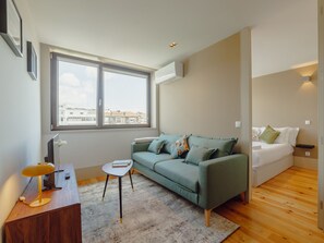 A modern living room with a green sofa, a small side table, a wooden desk with a lamp, and a large window offering a city view. - Apartment  by Interhome (Porto)