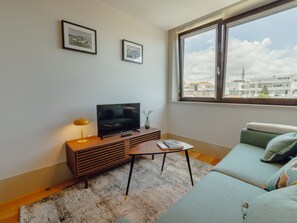 Ein modernes Wohnzimmer mit einem Fernseher auf einer Holzkonsole, einem kleinen Tisch mit Büchern, einer Couch mit Kissen und einem großen Fenster mit Blick auf die Stadt.