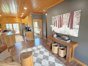 A spacious cabin interior with wooden floors, a kitchen area, a dining space, and a large window offering a view of the outdoors. - Reiser's Ridge Cabin (Custer)