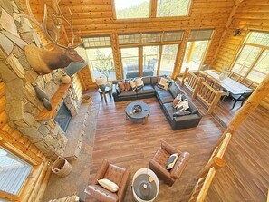 A log cabin interior with a stone fireplace, leather sofas, a dining area, and a large window overlooking the outdoors. - Reiser's Ridge Cabin (Custer)
