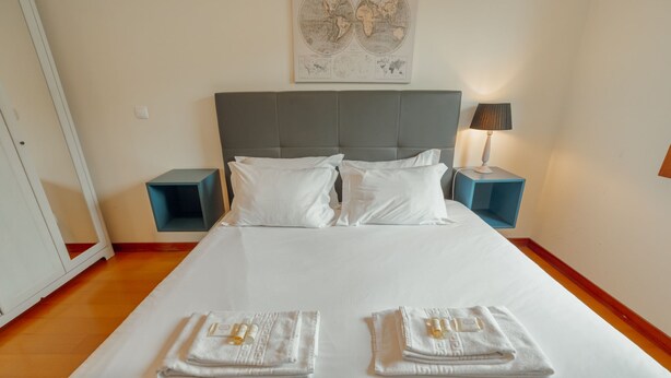 Uma cama bem arrumada com roupa de cama branca, duas mesas de cabeceira com abajures e um mapa-múndi na parede.