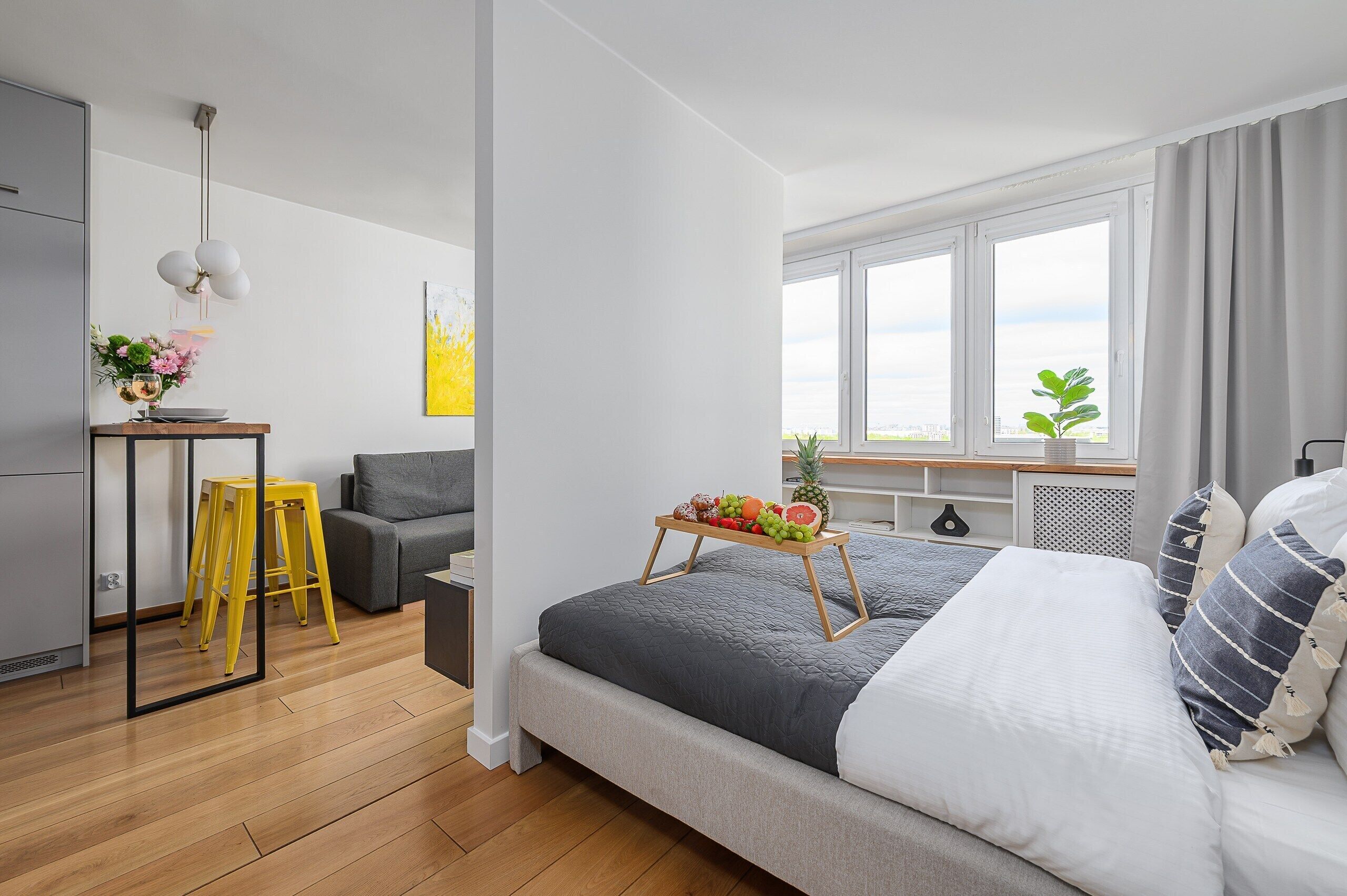 A modern bedroom with a bed, a small table with fruit, a grey sofa, and a window with a plant.