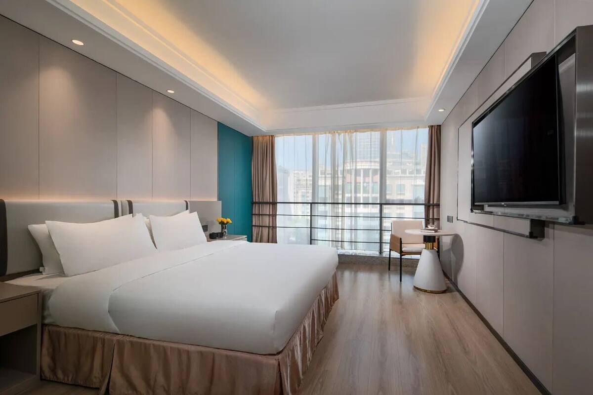 Una habitación de hotel moderna con una cama grande, un televisor de pantalla plana, una mesita pequeña y vistas de la ciudad a través de amplios ventanales.