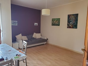 A room with a bed, a table with a tablecloth, a chair, and two paintings on the wall.