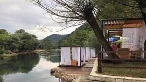 A riverside wooden deck with a small white structure, possibly a cabana or storage shed, and a covered seating area with a table and chairs.