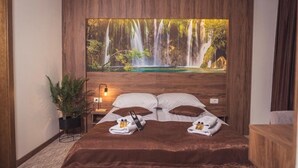 A bedroom with a large wall-mounted waterfall painting, a bed with brown bedding, a bedside table with a lamp, and a potted plant.