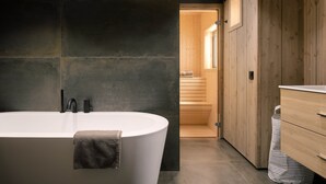 Une salle de bain moderne avec une baignoire sur pieds, des murs recouverts de carreaux foncés, des meubles en bois et un sauna visible.