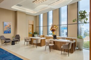 A modern hotel lobby with a reception desk, seating area, large windows, and a decorative chandelier.