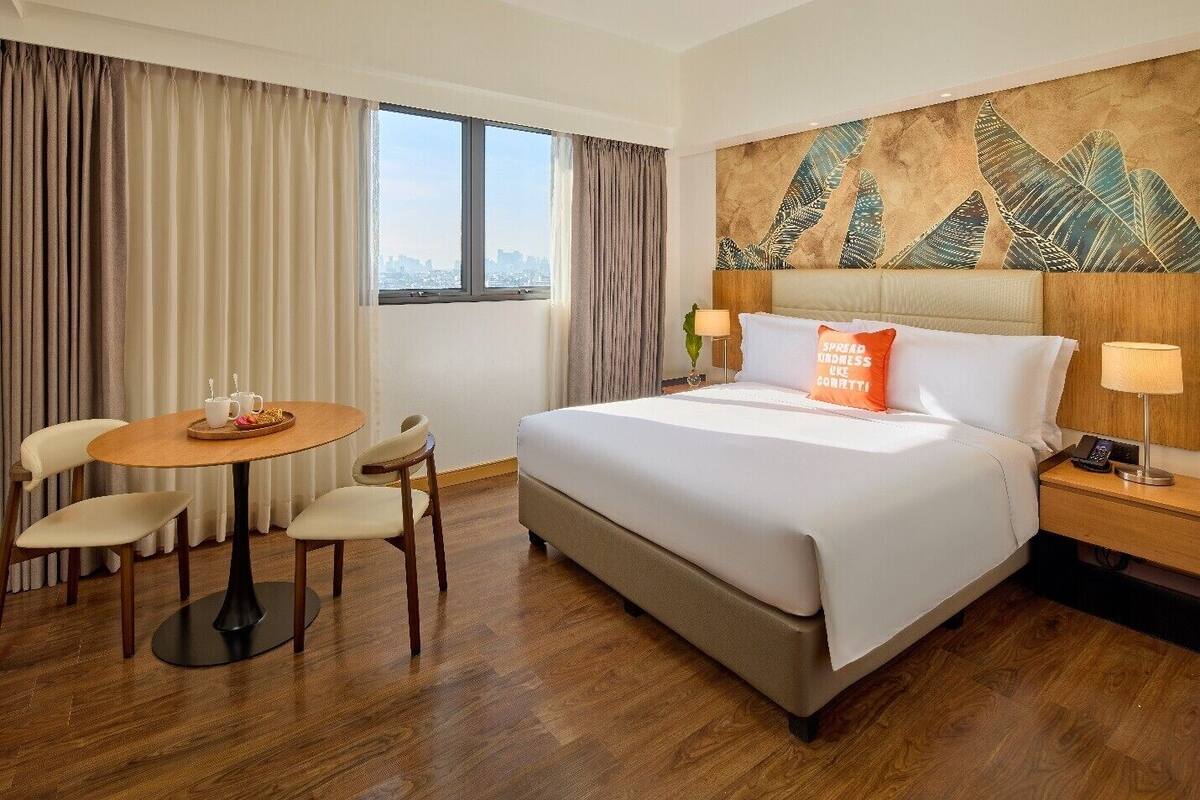Una habitació d'hotel amb un llit, una taula rodona amb cadires, una tauleta de nit i una finestra amb cortines.