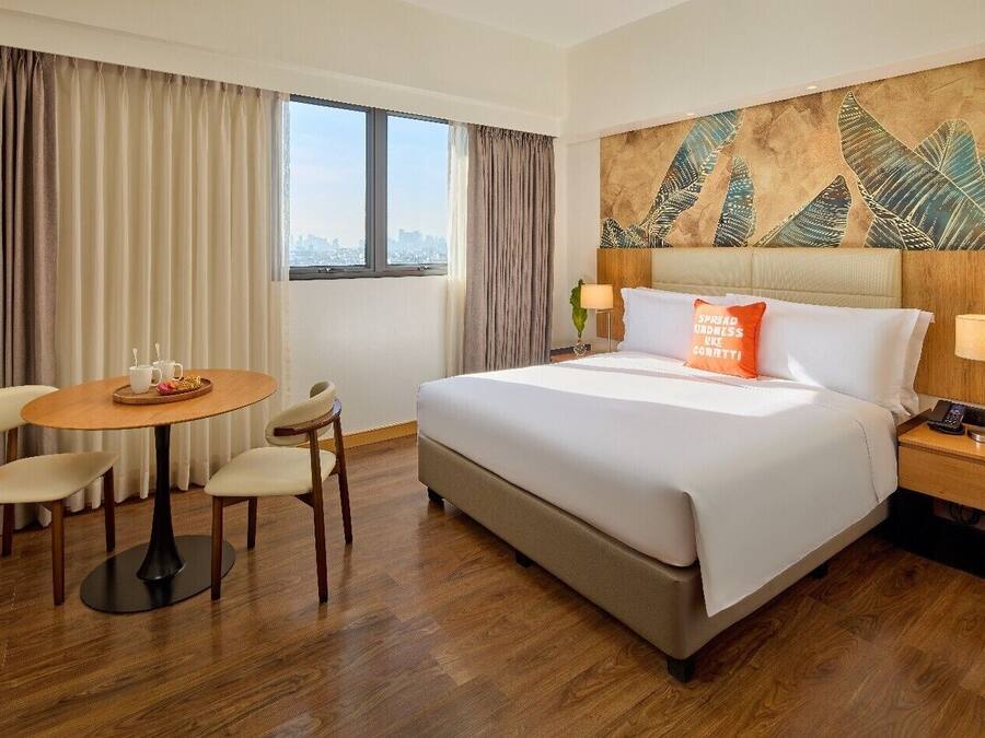 Quarto de hotel com cama, mesa redonda com cadeiras, mesa de cabeceira e janela com cortinas.
