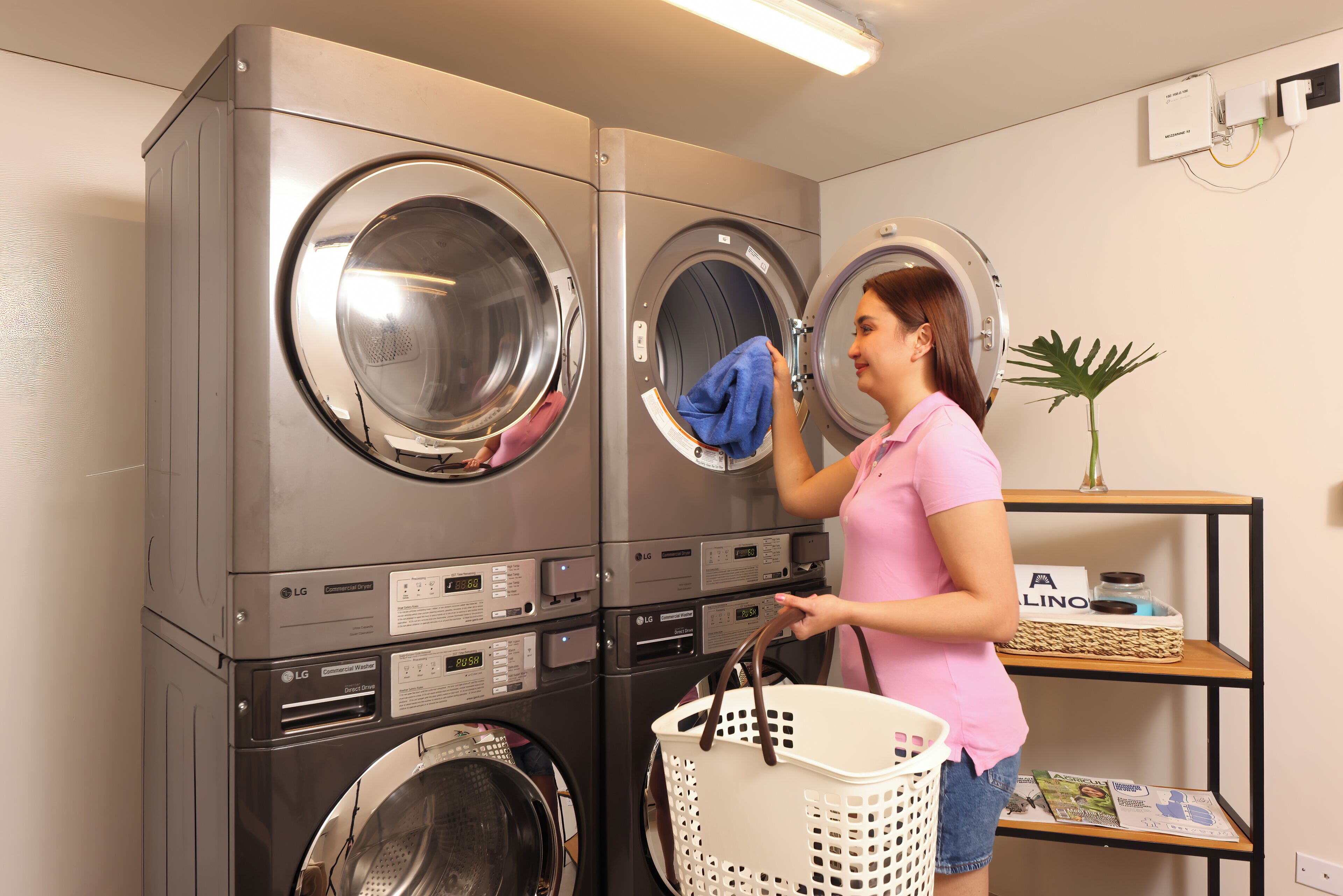 A laundry room with stacked washers and dryers, a basket, and a shelf with magazines.