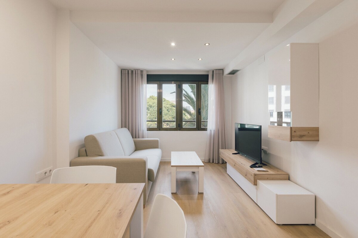 Uma sala de estar moderna com um sofá, uma televisão sobre um suporte de madeira, uma mesa de jantar e uma janela com cortinas.