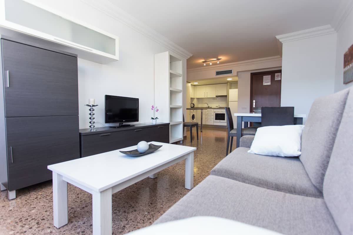 A modern living room with a sofa, a TV, a white table, and a kitchen in the background.