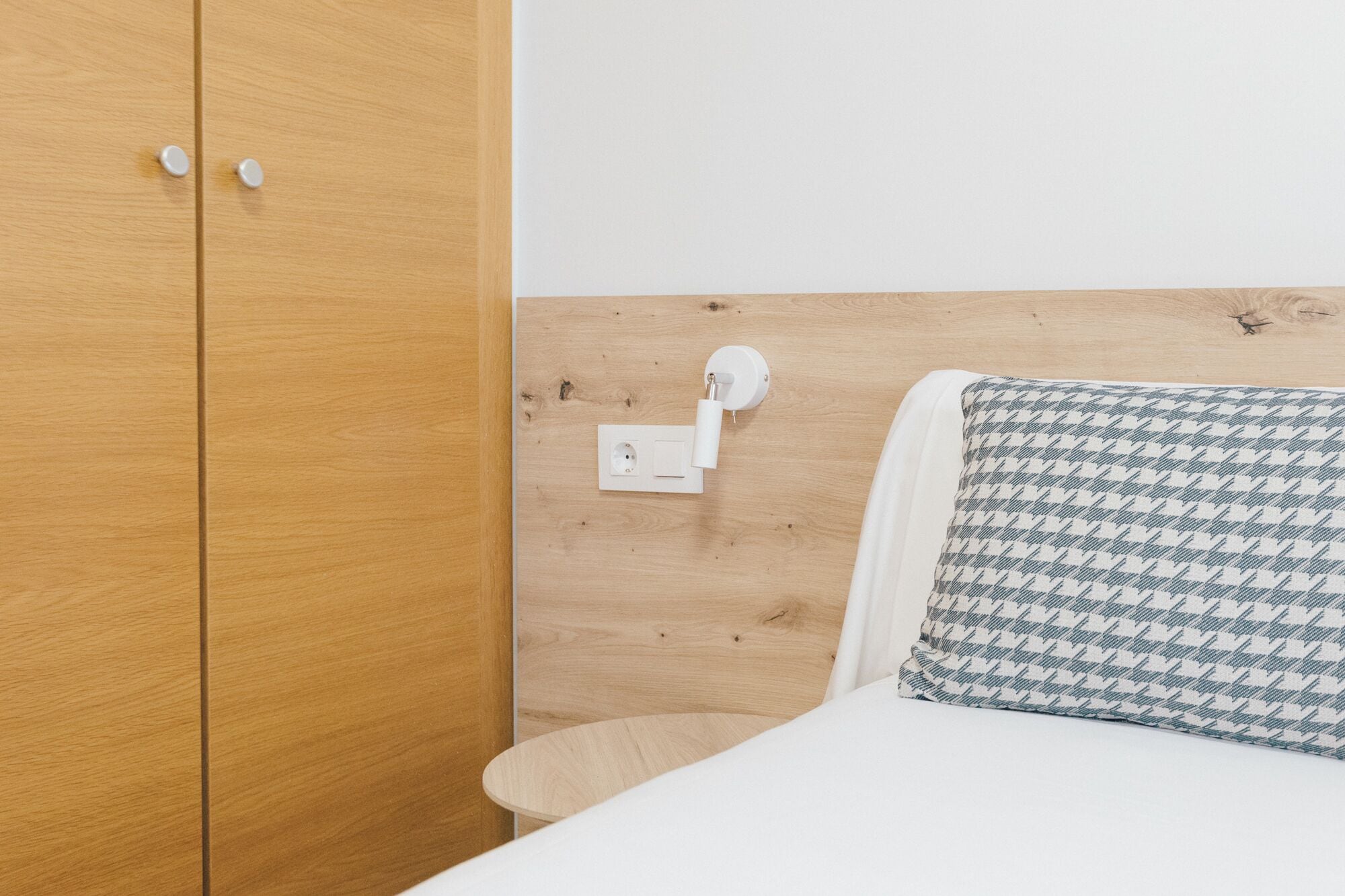 A wooden wardrobe with two doors, a white wall socket, and a bed with a patterned pillow.