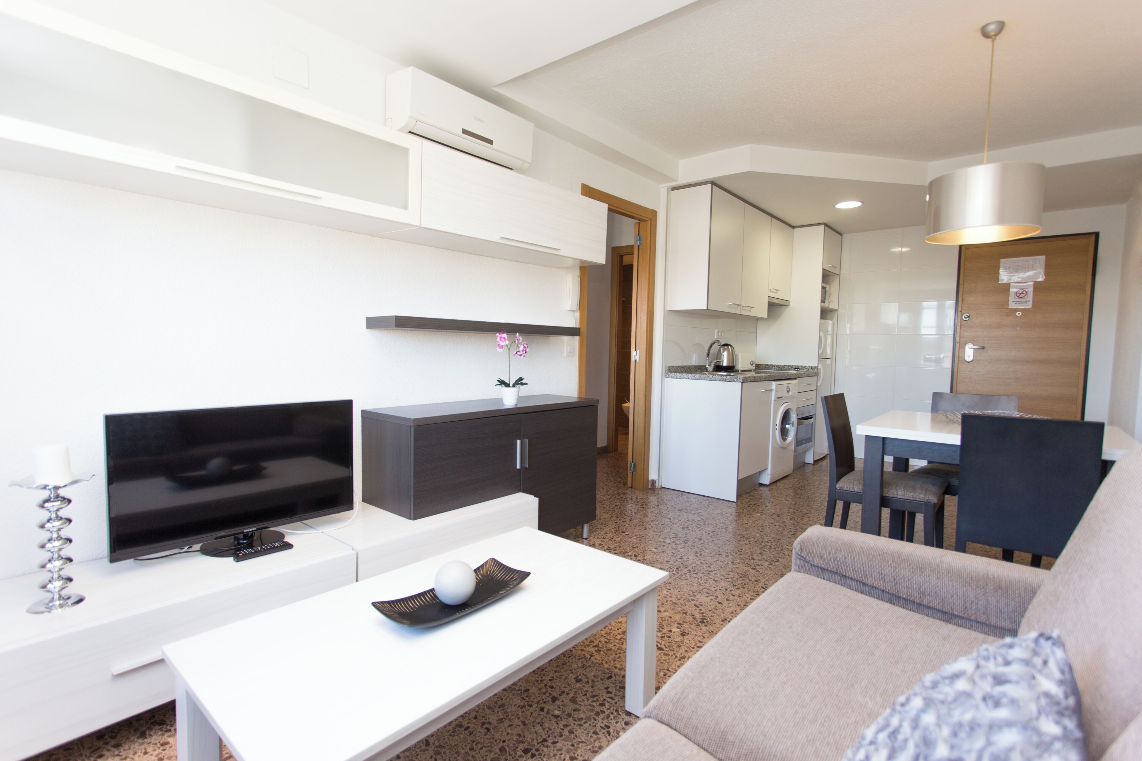 A modern living room with a flat-screen TV, a white coffee table, a sofa, and a kitchen area in the background.