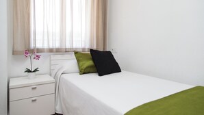 A single bed with a white mattress and two pillows, a bedside table with a plant, and a window with sheer curtains. - Monlar Campanar (Valencia)