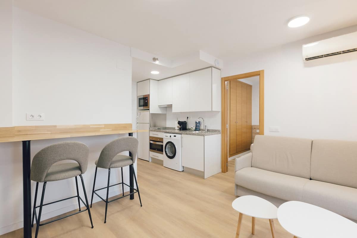 A compact kitchen with a washing machine, a microwave, and a coffee maker. There are two bar stools and a beige sofa with two small white tables.