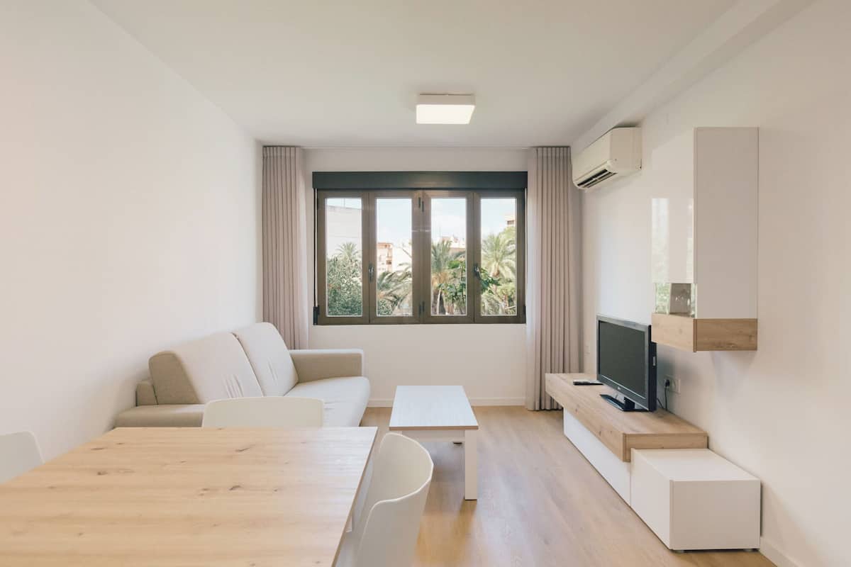 A modern living room with a sofa, a dining table, a TV, and a large window with a view of buildings and trees.