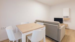 A minimalist living room with a wooden table, two white chairs, a grey sofa, and a TV on a wooden stand. - Monlar Campanar (Valencia)