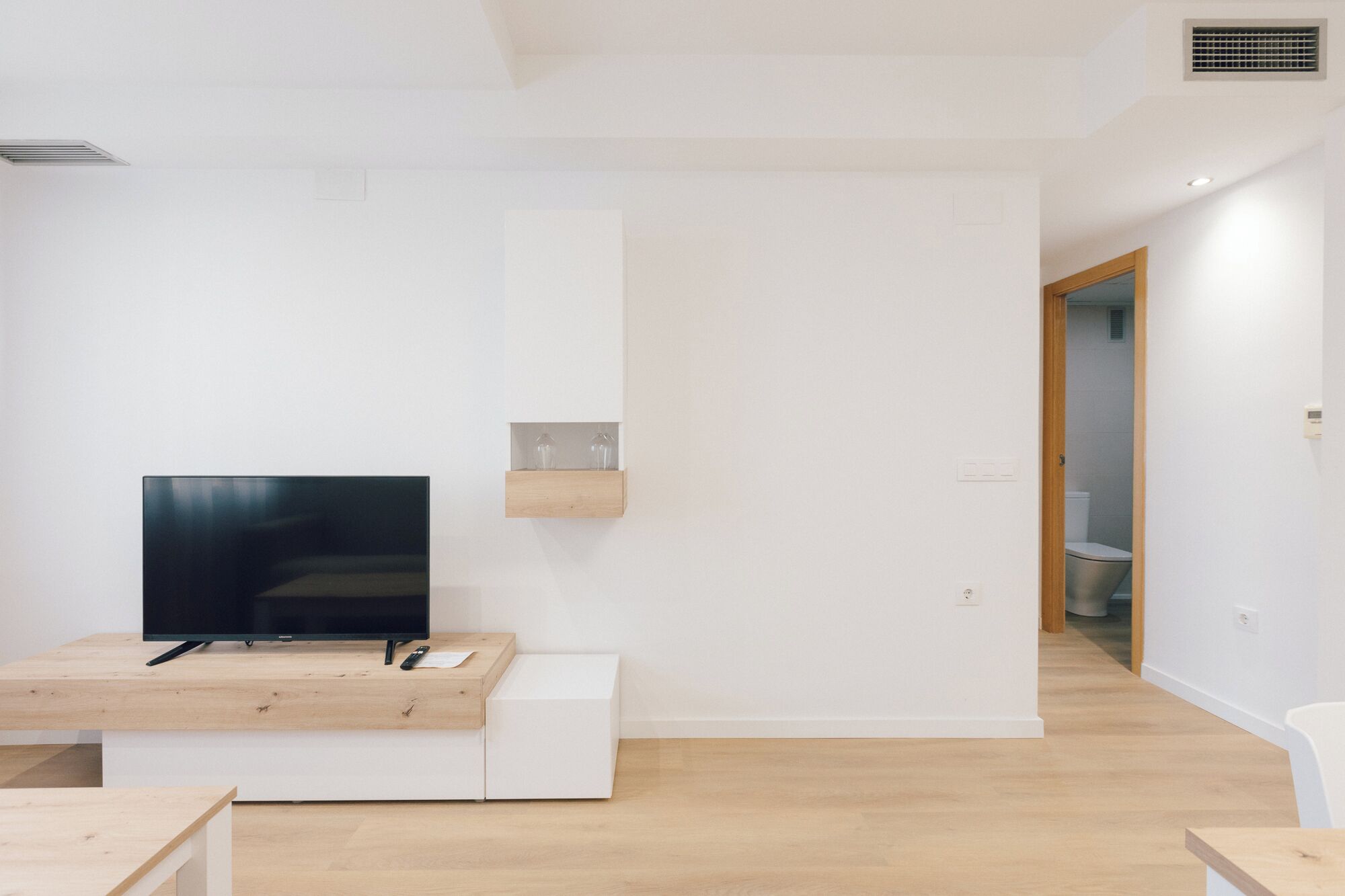A modern living room with a flat-screen TV on a wooden table, a built-in shelf with glassware, and a visible bathroom door.