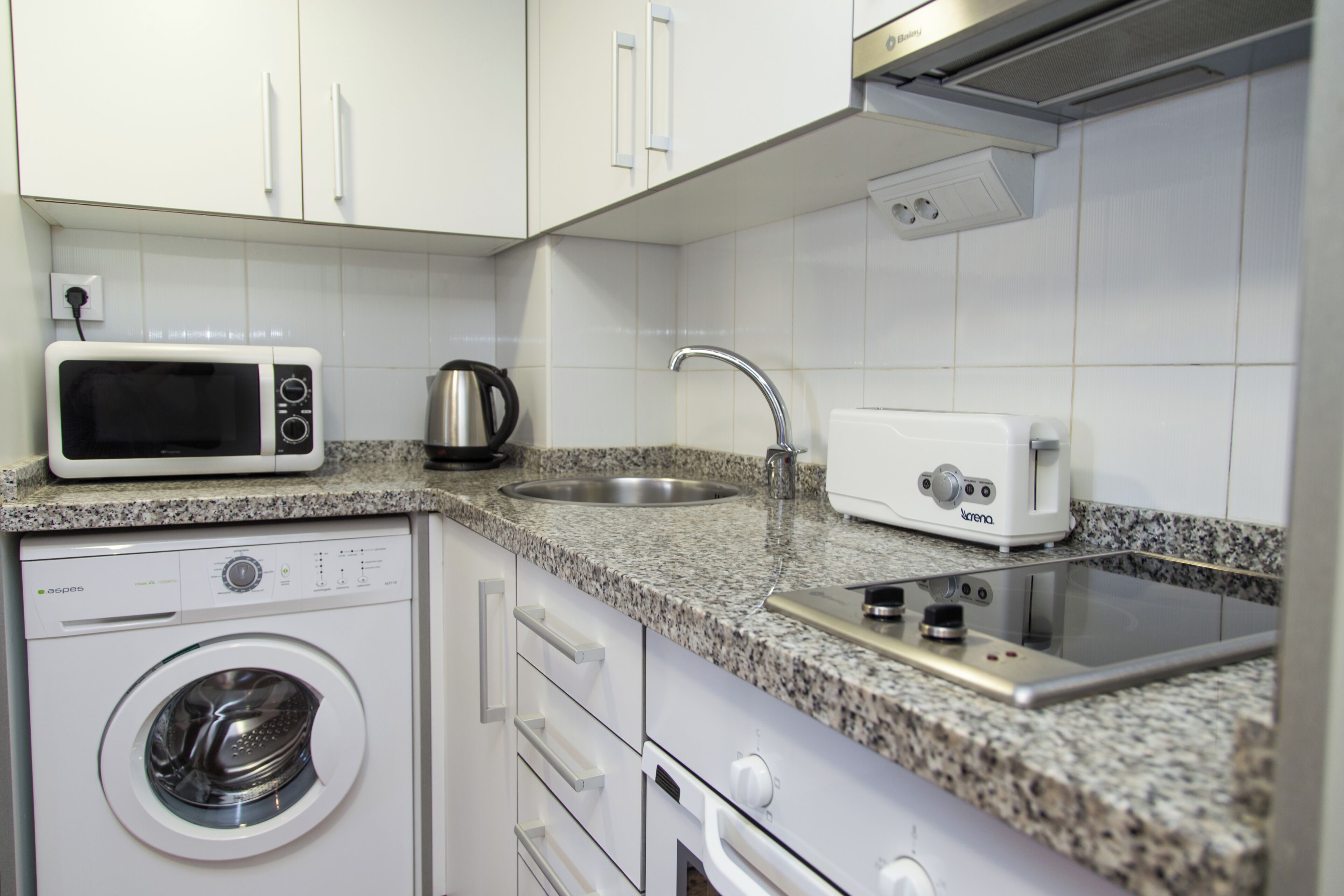 A compact kitchen with a washing machine, microwave, toaster, and stainless steel sink.
