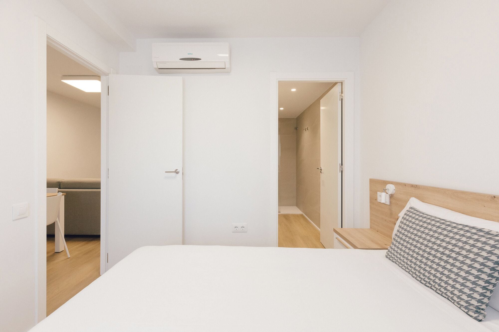 A bedroom with a bed, a bedside table, a nightstand, a wooden headboard, a wall-mounted air conditioner, and a door leading to a bathroom.