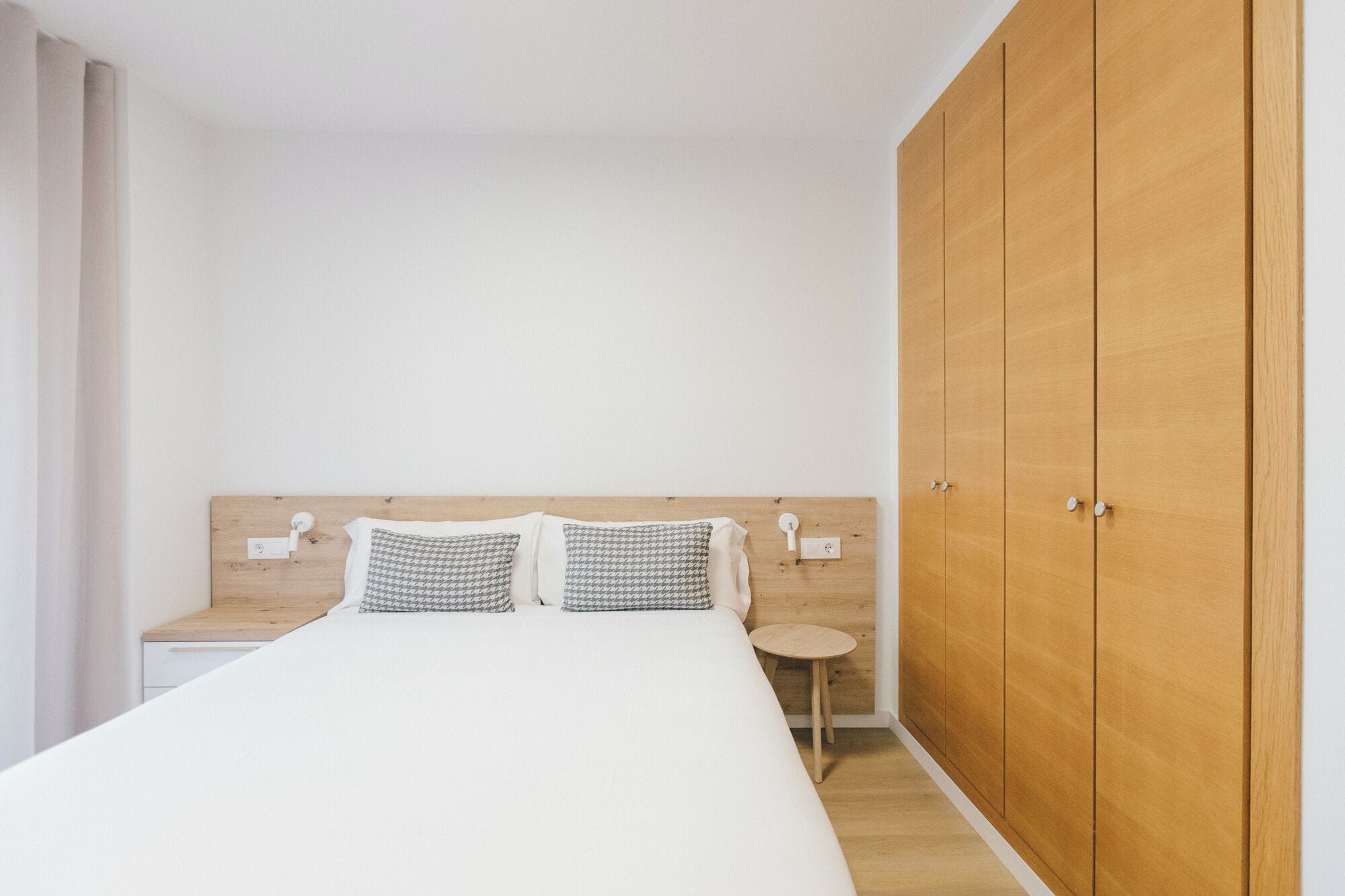 Quarto com guarda-roupa de madeira, cama com almofadas estampadas, mesa de cabeceira e uma pequena mesa redonda.