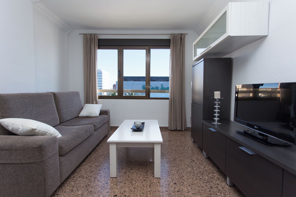 Quarto de hotel moderno com sofá, uma mesa pequena, televisão e vista para a cidade.
