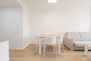 A minimalist living room with a wooden dining table and white chairs, a beige sofa, and a small white side table. - Monlar Campanar (Valencia)