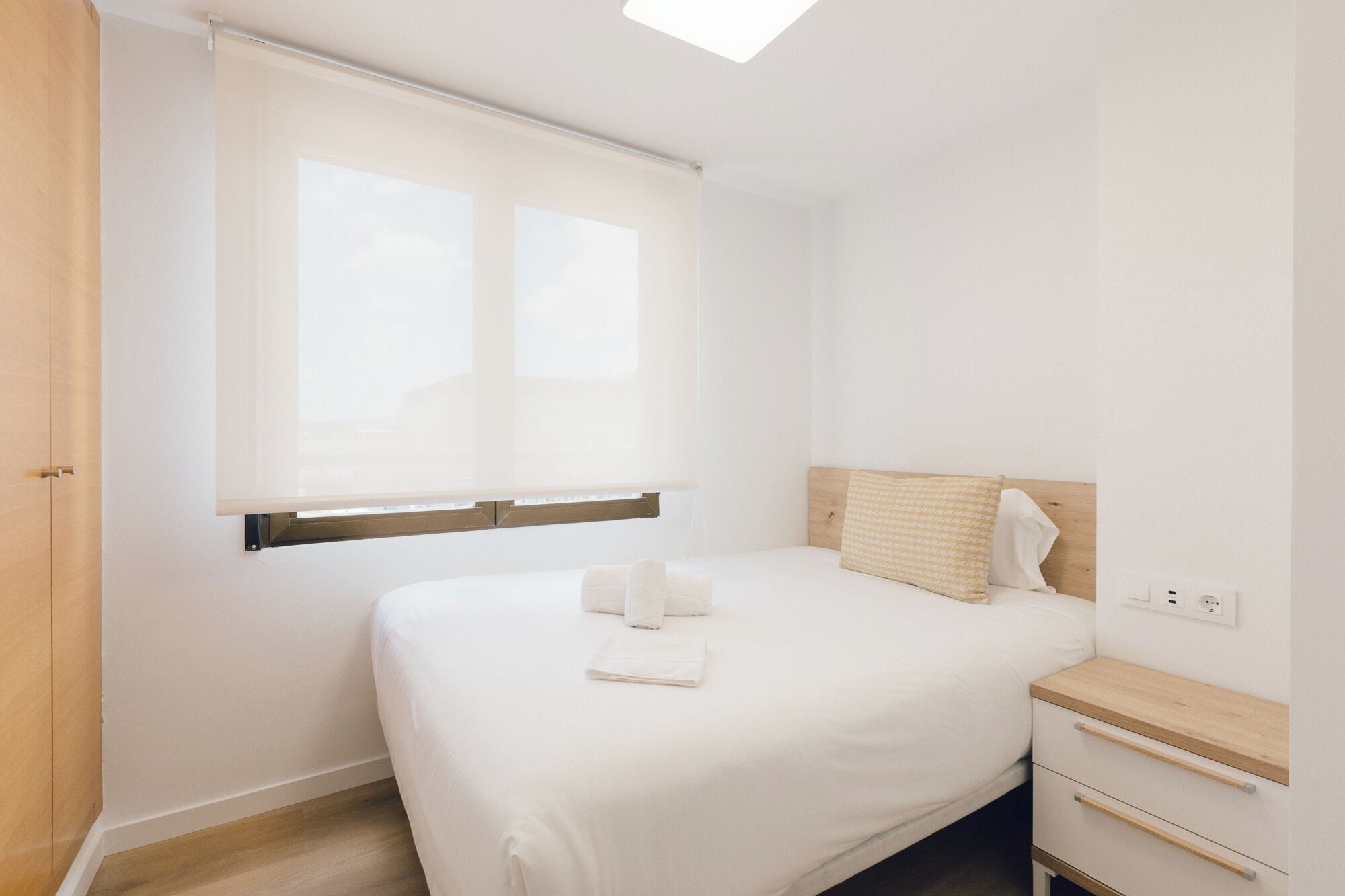 Une chambre d’hôtel au style minimaliste, avec un grand lit, une table de chevet en bois et une fenêtre équipée de stores.