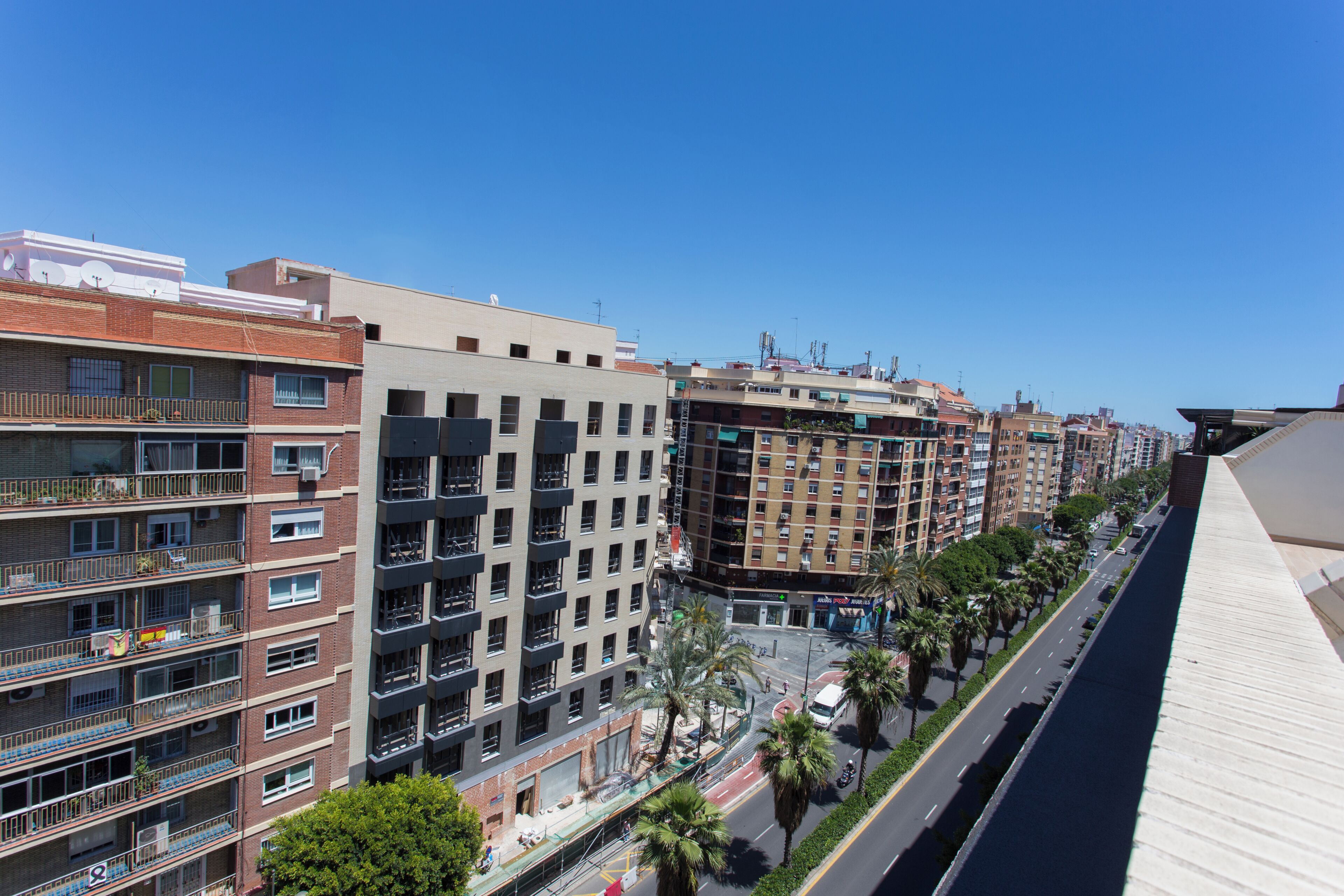 A multi-story building with balconies, a street with palm trees, and a clear blue sky.