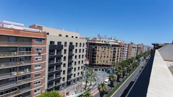 A multi-story building with balconies, a street with palm trees, and a clear blue sky.