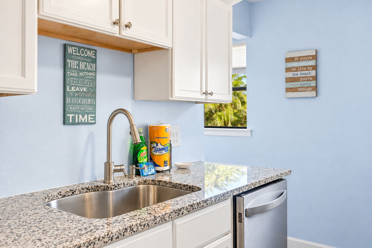 A kitchen with a granite countertop, stainless steel sink, and white cabinets. There are two wall-mounted signs, one with a beach message and another with a motivational quote.
