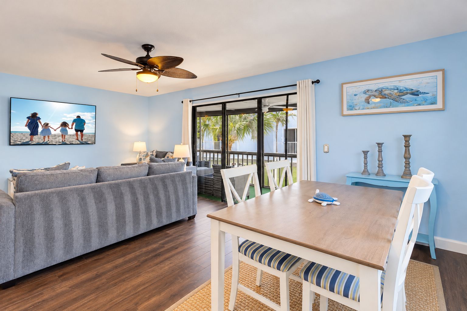 A room with a sofa, a dining table, chairs, a ceiling fan, and a large window with a view of palm trees.