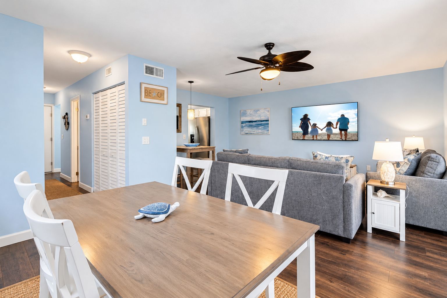 Un salon avec un canapé gris, une table à manger avec des chaises, un ventilateur de plafond et, au fond, un coin cuisine.
