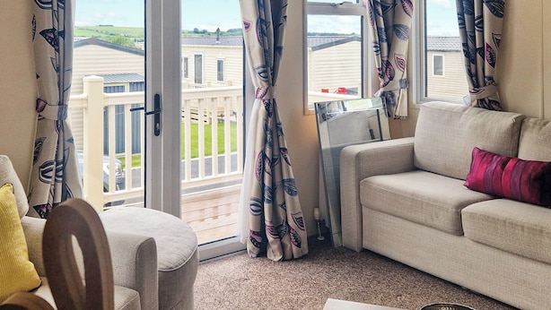 A living room with a sofa, armchair, and a dining table with chairs. There is a sliding glass door leading to a balcony with a view of houses. - Birch 3-bedroom Pet Friendly (Clynderwen)