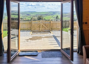 Une pièce dotée d’une grande porte-fenêtre coulissante donnant sur un patio extérieur avec une porte en bois, un chemin en pierre et une vue sur des champs verdoyants.