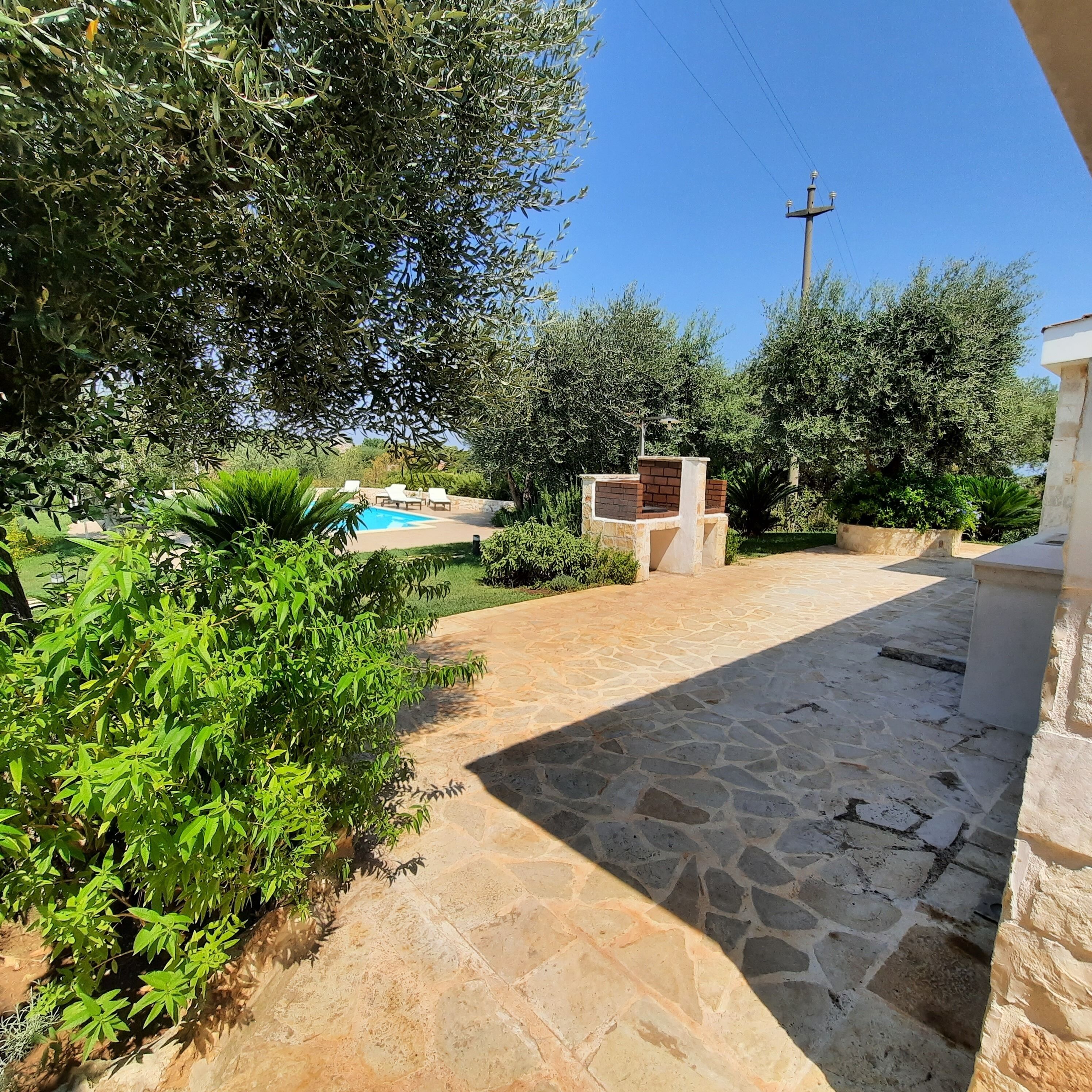 A stone pathway leading to a pool area surrounded by lush greenery and a brick barbecue.