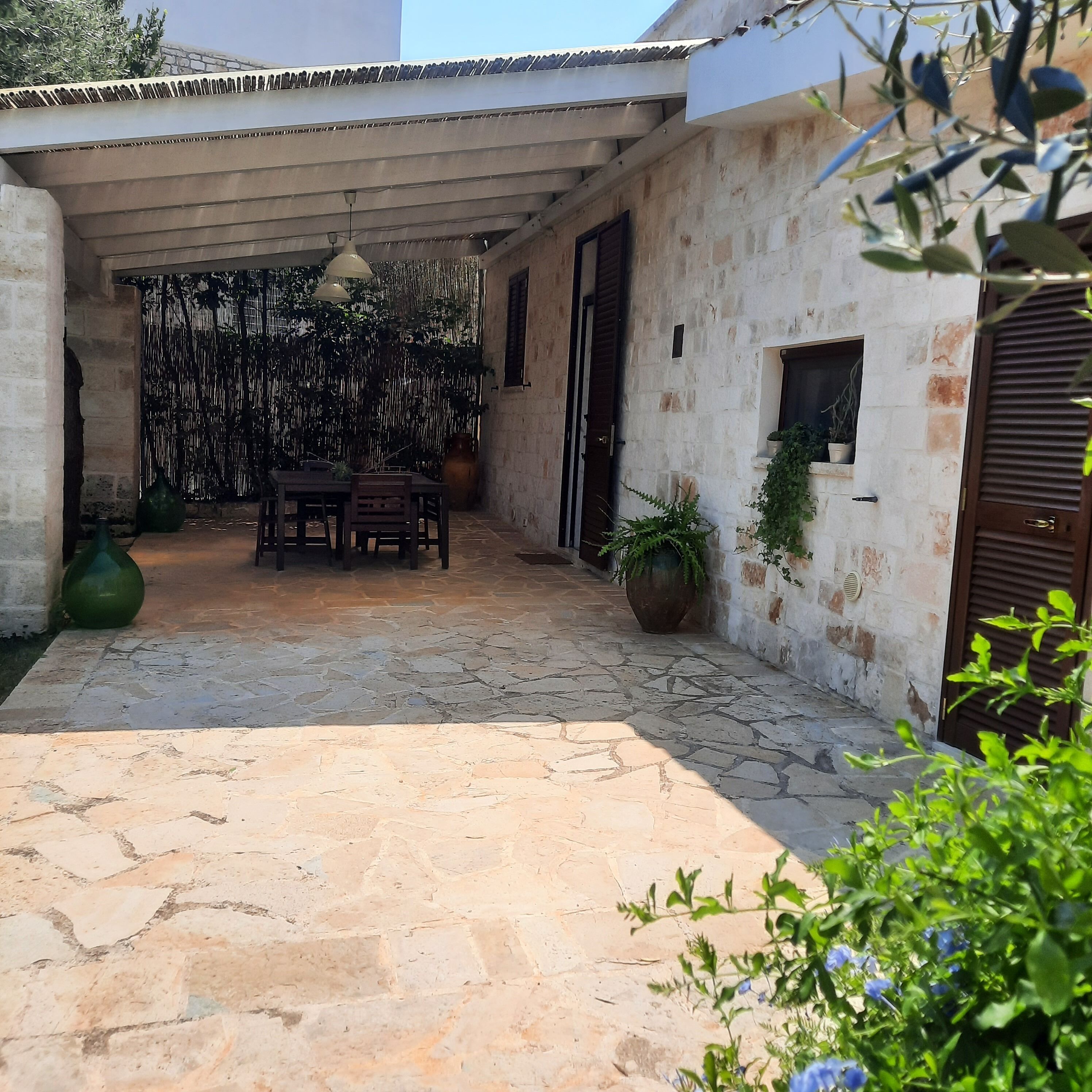 Ein überdachter Patio-Bereich mit Steinboden, einem Tisch und Stühlen sowie Topfpflanzen.