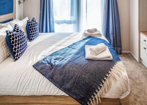 Un lit bien fait, recouvert d’une couverture bleue et blanche, avec une serviette enroulée et des coussins à motifs.