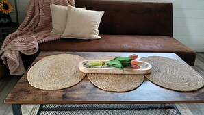 Un canapé marron agrémenté d’une couverture tissée et de coussins, une table en bois avec des sets de table tissés et un plateau de fleurs.
