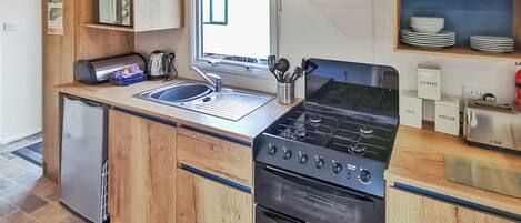 A compact kitchen with a built-in stove, oven, and sink. There are wooden cabinets, a small refrigerator, and a window with a view.