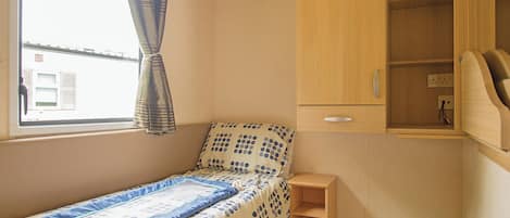 A small bedroom with a single bed, a wooden wardrobe, a small shelf, and a window with curtains.