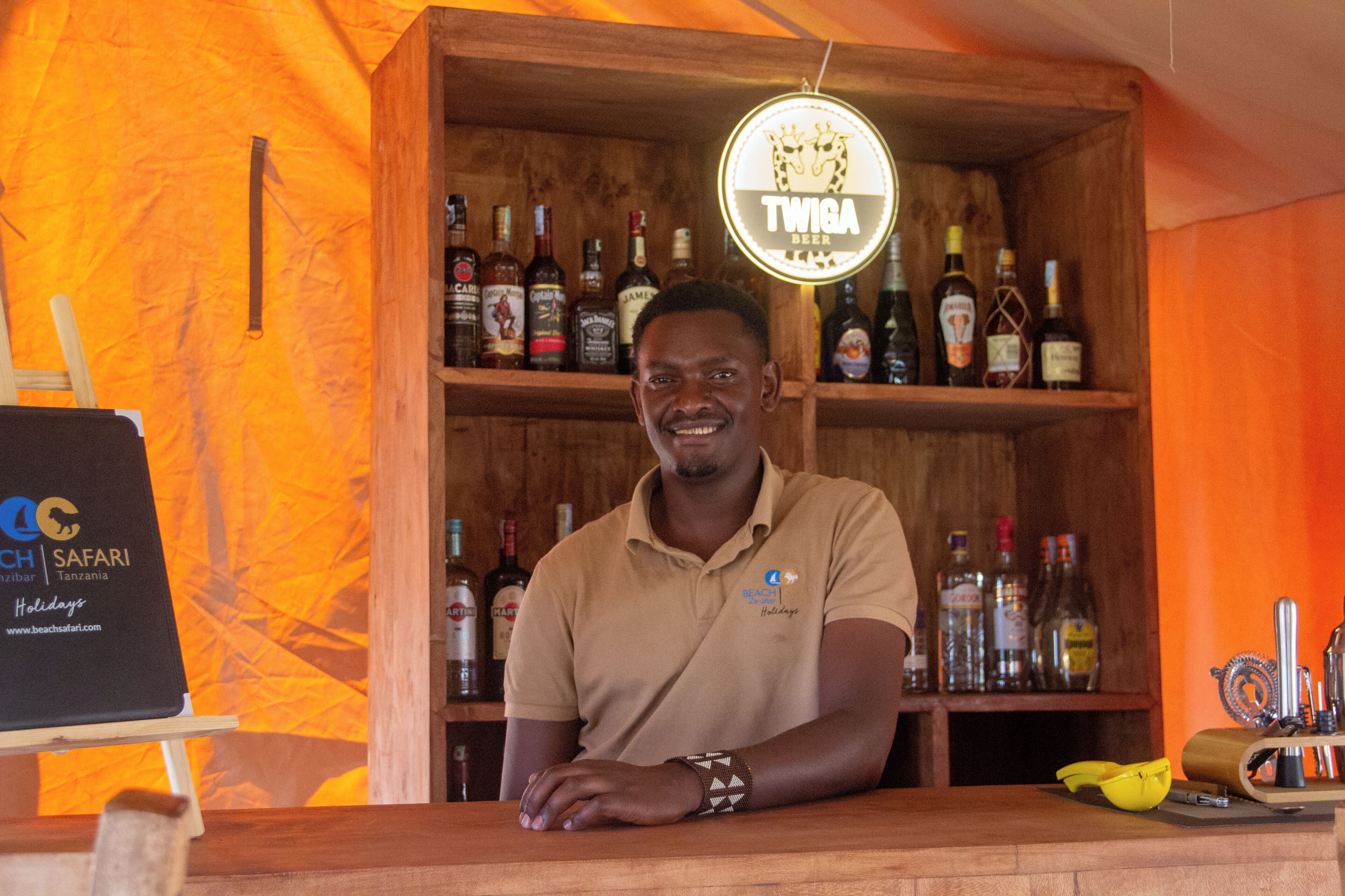 Un bar con un estante de madera repleto de botellas variadas, un letrero que indica 'TWIGA BEER' y el logotipo de 'BEACH Safari Tanzania' a la izquierda.