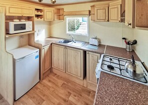 A compact kitchen with wooden cabinets, a microwave, a sink, a toaster, and a gas stove. - Bronze Plus 3 Bed Pets Sleeps 6 (Clarach Bay, Nr Aberystwyth)