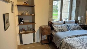 A bedroom with a bed, a nightstand, a dresser, a window with a view, and shelves with various items.