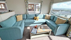 Ein modernes Wohnzimmer mit einem salbeengrünfarbenen Ecksofa, einem Couchtisch aus Holz mit Büchern und einer Teekanne sowie großen Fenstern mit Blick auf die Landschaft.